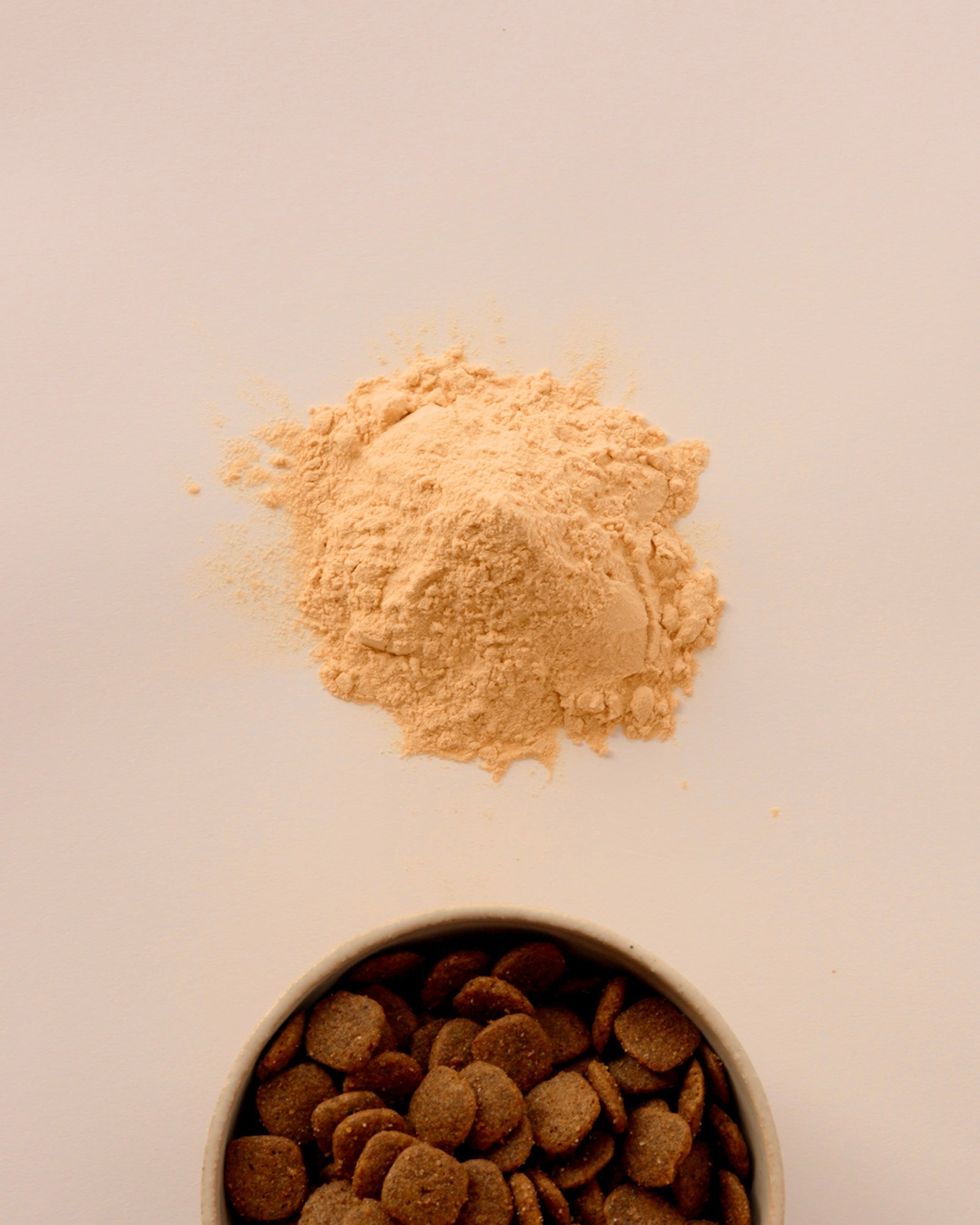Pile of yellow powder above a bowl of brown pet food on a light background